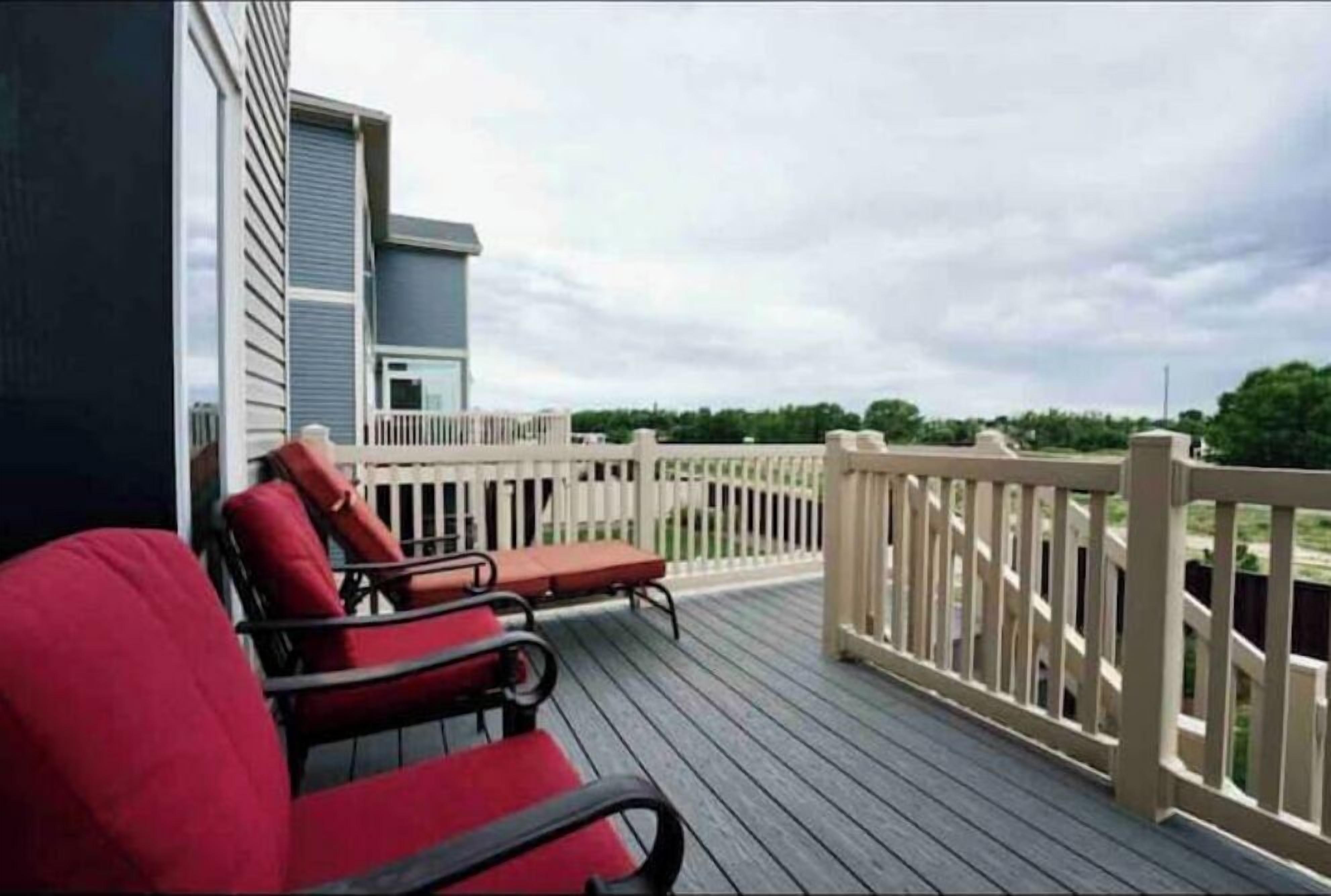 A wooden deck with two red chairs and a table, a white railing, and a building in the background.