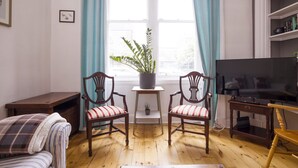 A living room with a wooden floor, two wooden chairs with striped cushions, a small table with a potted plant, a television, and a bookshelf. - Splendid Apartment near Haymarket (Edinburgh)