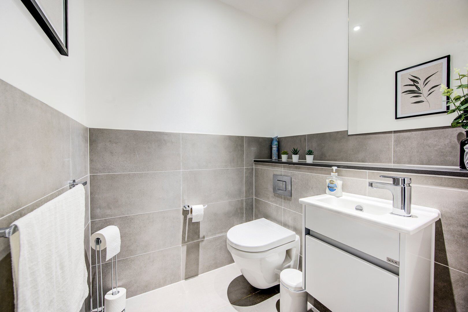 Une salle de bain moderne avec des carreaux gris, des toilettes blanches, un lavabo avec un robinet chromé et une petite étagère contenant des articles de toilette.