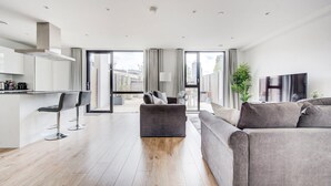 Ein modernes Wohnzimmer mit grauem Sofa, einem Couchtisch und einem Fernseher. Es gibt eine Küchenzeile mit weißen Schränken und einer Bar.
