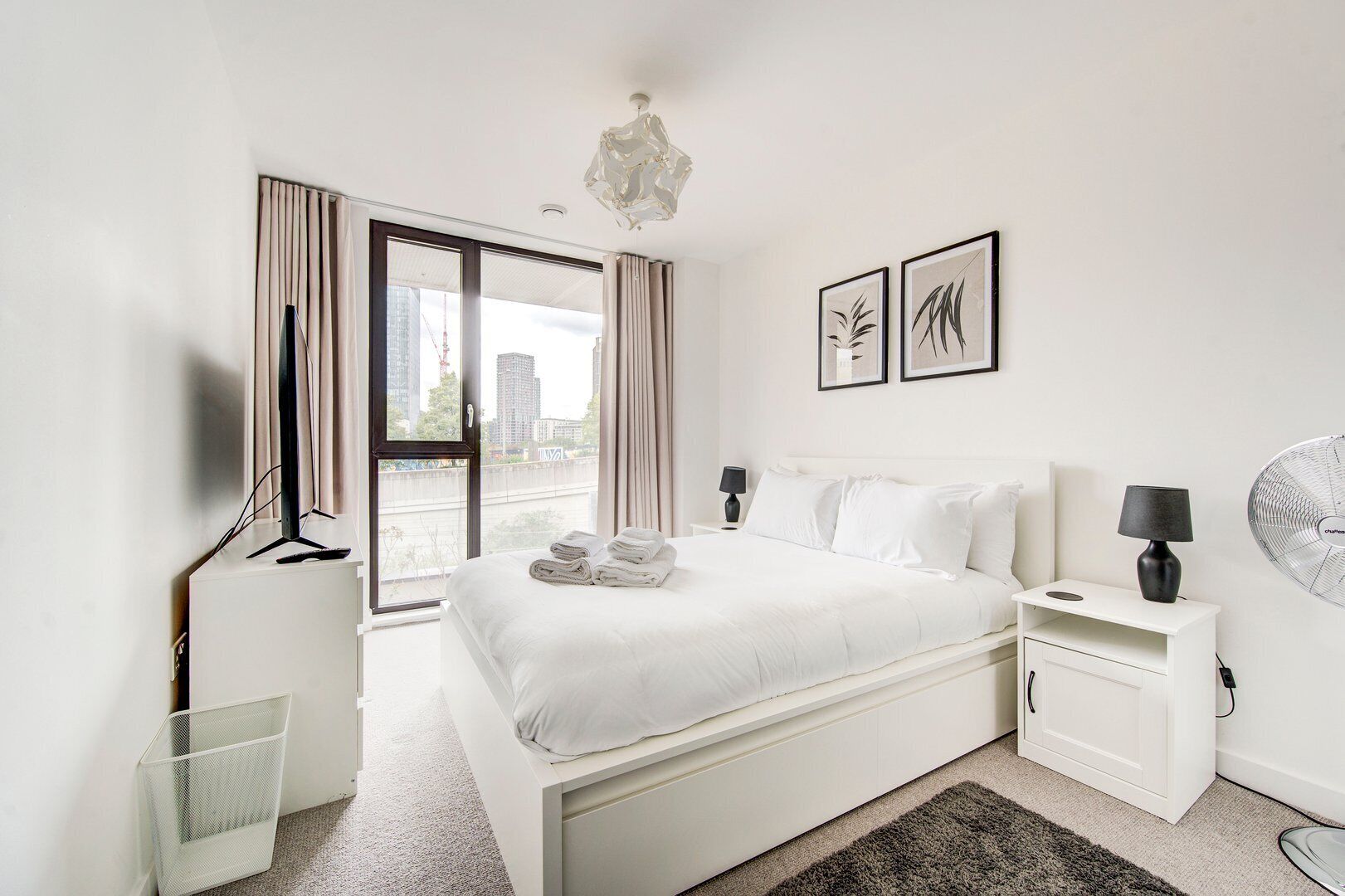 Une chambre d’hôtel moderne, dotée d’un grand lit, d’un bureau, d’une chaise, d’un ventilateur sur pied et offrant une vue sur la ville par la fenêtre.