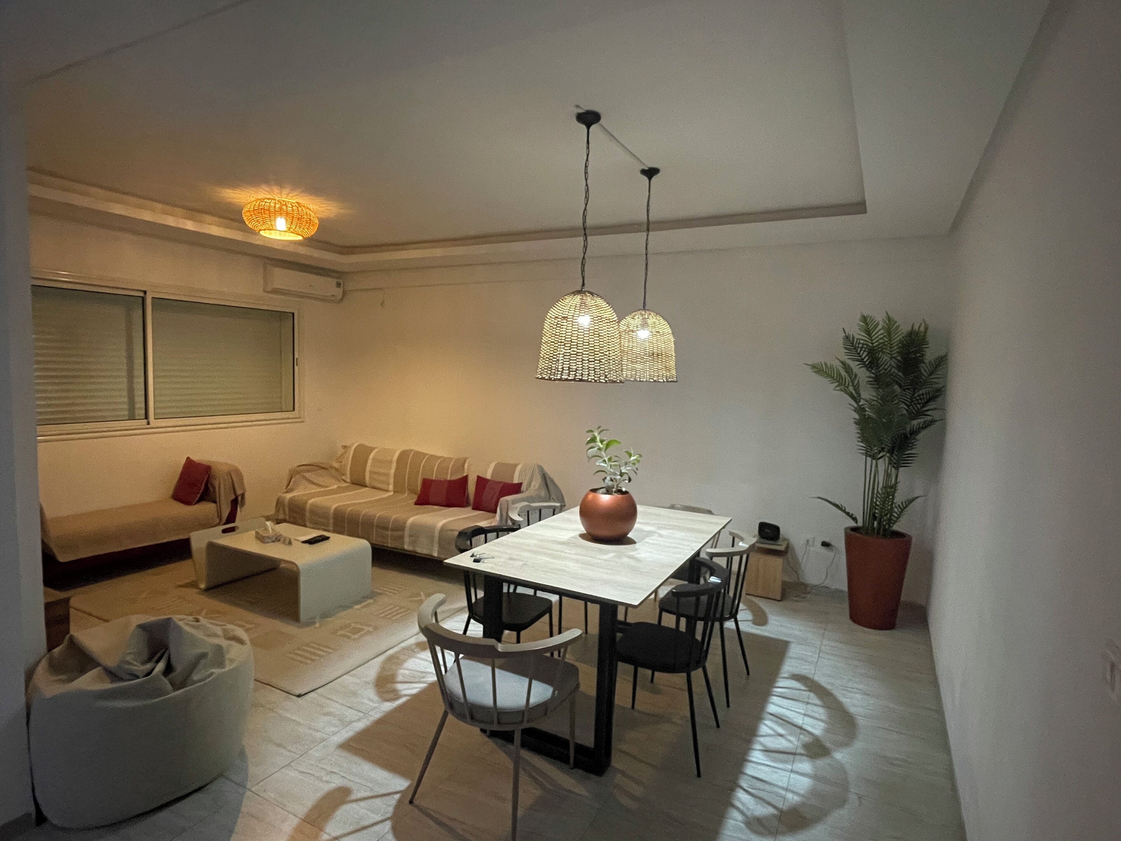Un salon moderne comprenant un canapé, une table à manger, des chaises et une table basse. On y trouve deux lampes suspendues et une plante en pot.