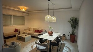 A modern living room with a sofa, a dining table, chairs, and a coffee table. There are two hanging lights and a potted plant. - Appartement S1 Avec Grande Terrasse à 10 min de la Plage (Mrezga)