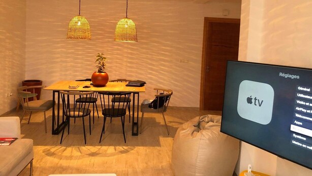 A modern living room with a dining area, featuring a large flat-screen TV displaying the Apple TV interface. - Appartement S1 Avec Grande Terrasse à 10 min de la Plage (Mrezga)