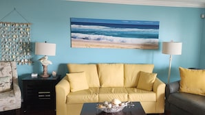 Uma sala de estar com um sofá amarelo, uma mesa de centro preta com objetos decorativos, um quadro com vista para o oceano emoldurado e uma luminária em uma mesinha lateral.