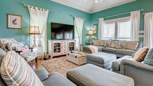 A living room with teal walls, a ceiling fan, a flat-screen TV, and a large window with white curtains.
