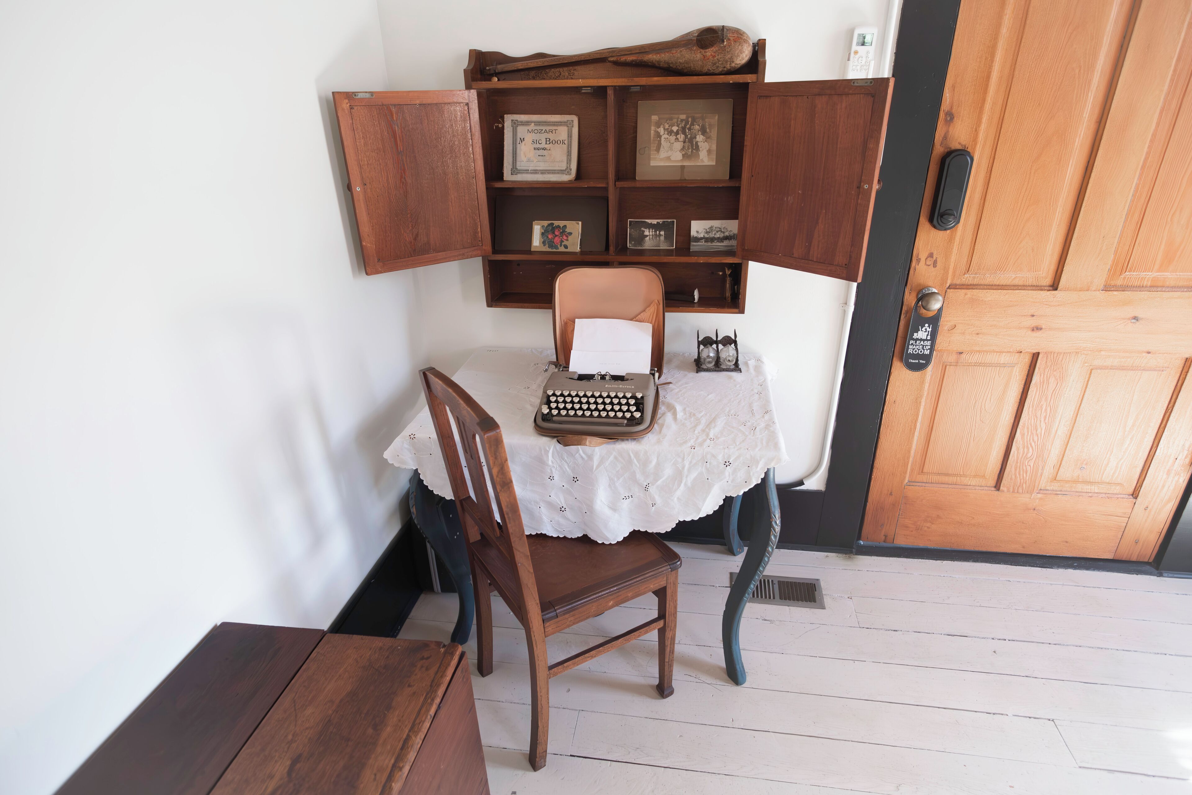 A wooden chair with a typewriter on a lace table, a wooden cabinet with framed items, and a wooden door.