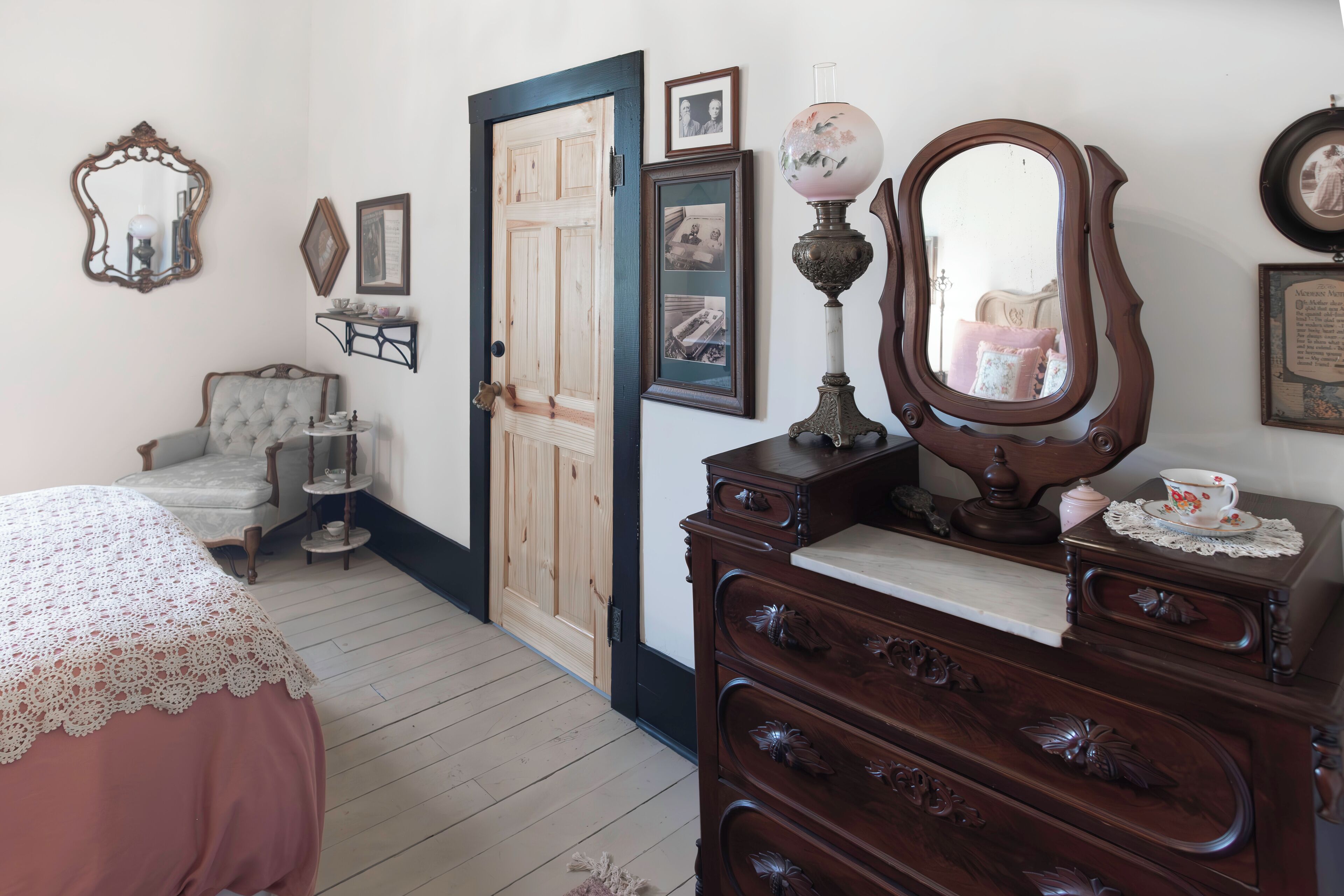 A room with a wooden dresser, a chair, a bed with a lace cover, and a door leading to another room.