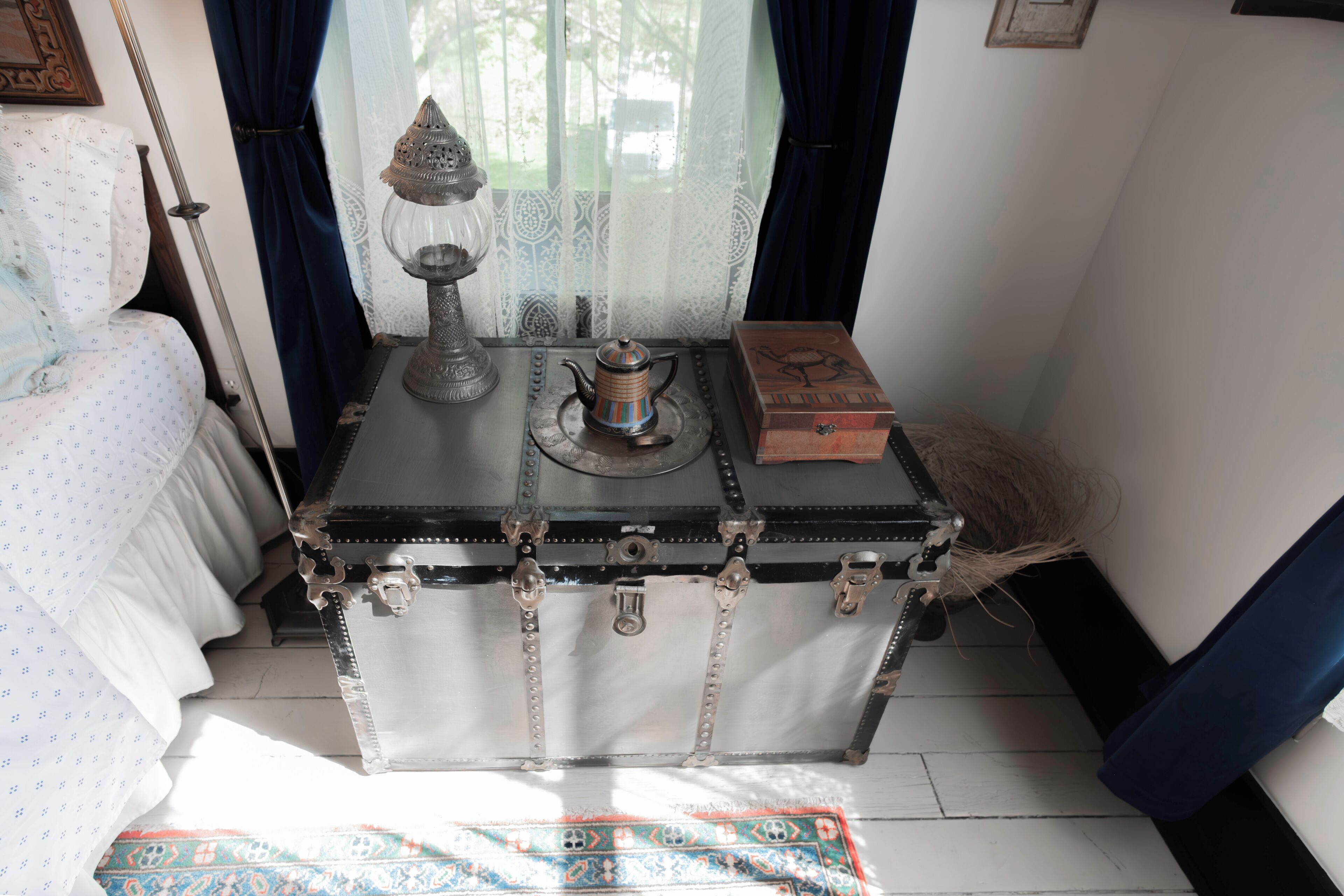 A room with a bed, a table with a lamp, a box, and a suitcase.