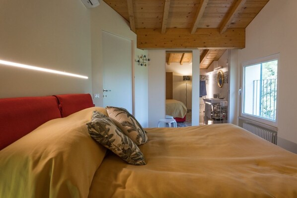 A bedroom with a bed, a red headboard, a wooden ceiling, a window, and a bathroom visible through an open door.