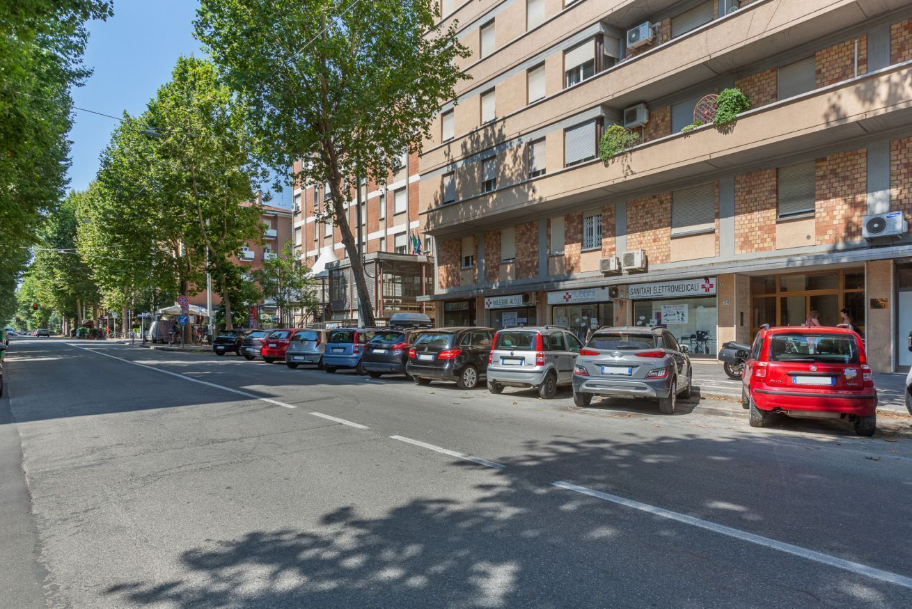 Una strada con auto parcheggiate, un edificio con il cartello 'SANTARI ELETROMEDICO' e alberi che fiancheggiano la carreggiata.