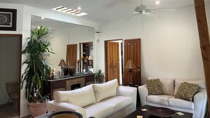 Un salón con un sofá blanco, un sillón blanco más pequeño, una mesa de centro, un espejo grande, un ventilador de techo y una planta en maceta.