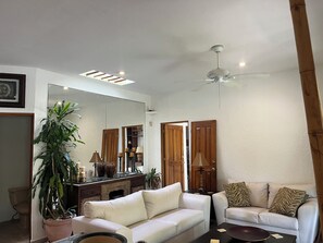 Ein Wohnzimmer mit einem weißen Sofa, einem kleinen weißen Sessel, einem Couchtisch, einem großen Spiegel, einem Deckenventilator und einer Topfpflanze.