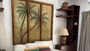 Una cama bien hecha con almohadones estampados, un cuadro colgado en la pared con palmeras, una lámpara de noche y una mesita de madera con una cesta.