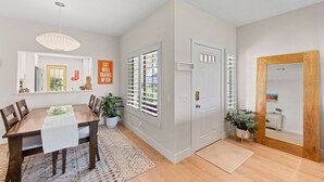 A dining area with a wooden table, chairs, and a white table runner. There are potted plants and a window with blinds. - Newly upgraded throughout and GORGEOUS sunset and mountain views! (Palm Desert)