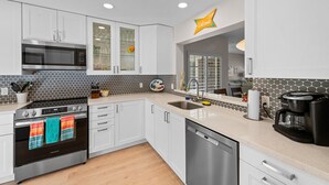 A modern kitchen with white cabinets, a stainless steel microwave, a coffee maker, and a dishwasher. - Newly upgraded throughout and GORGEOUS sunset and mountain views! (Palm Desert)