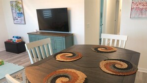 Una mesa de comedor con cuatro individuales de estampado espiral, sillas blancas, un televisor sobre un mueble y un cuadro en la pared.