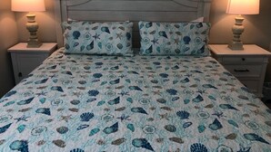 Una cama con colcha estampada de motivos florales y conchas marinas, flanqueada por dos mesitas de noche con lámparas, en una habitación con cabecera sencilla y curvada.
