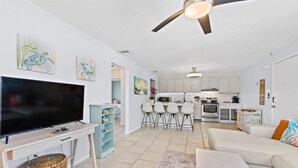 A modern living room with a flat-screen TV, a coffee table, and a sofa. There is a kitchen area with white cabinets and a dining table with four chairs. - Cape Canaveral Condo w/Pool, Steps to Beach Access (Cape Canaveral)
