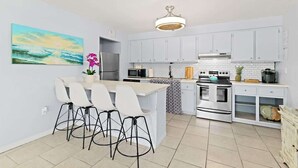 A modern kitchen with white cabinets, a central island with bar stools, stainless steel appliances, and a ceiling light fixture. - Cape Canaveral Condo w/Pool, Steps to Beach Access (Cape Canaveral)