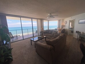 Ein Wohnzimmer mit einem großen Fenster und Blick auf einen Balkon, der auf das Meer hinausführt. Es gibt einen Deckenventilator, einen Couchtisch und eine Couch.