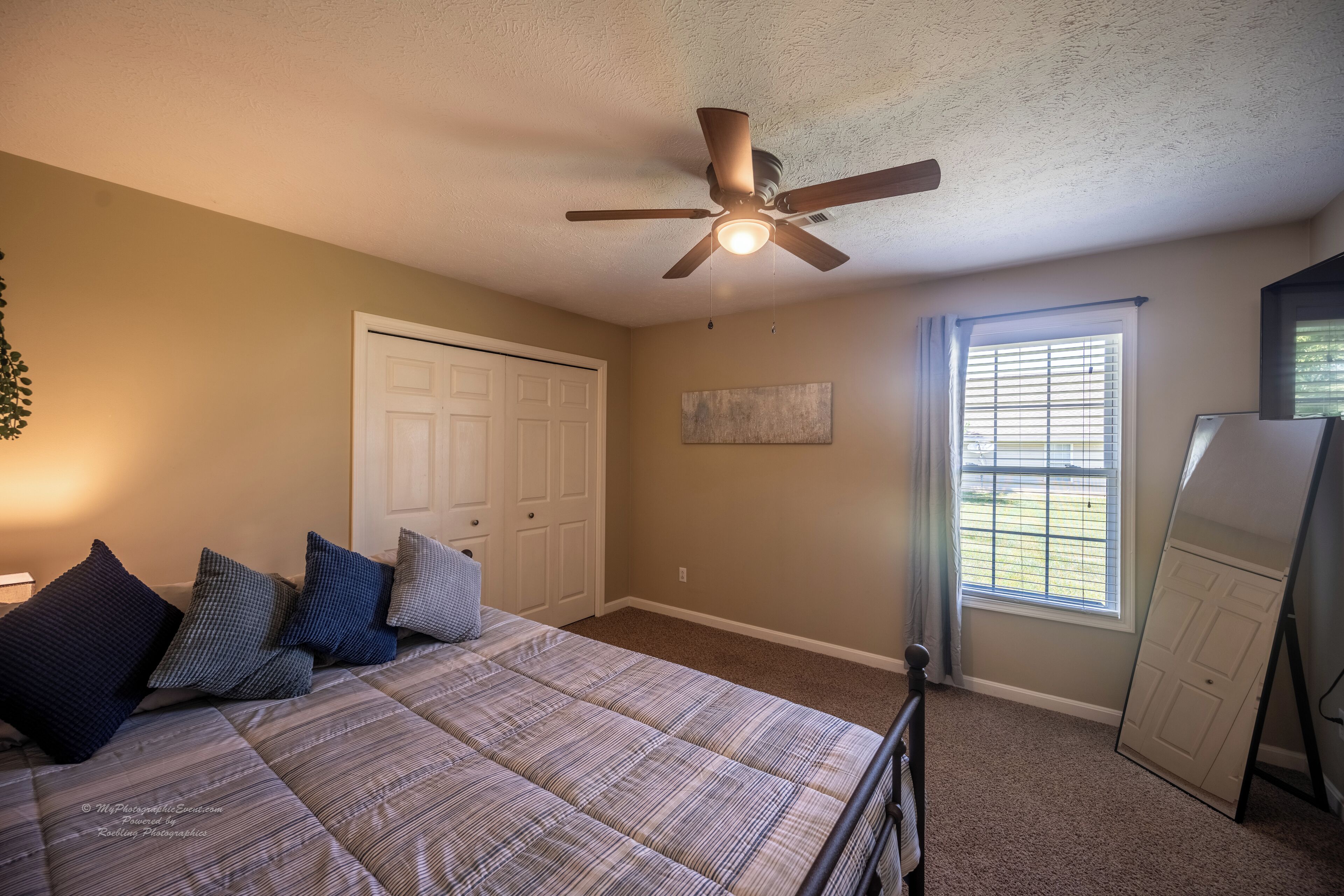 Une chambre à coucher avec un lit, un ventilateur de plafond, une fenêtre avec des stores et une porte.