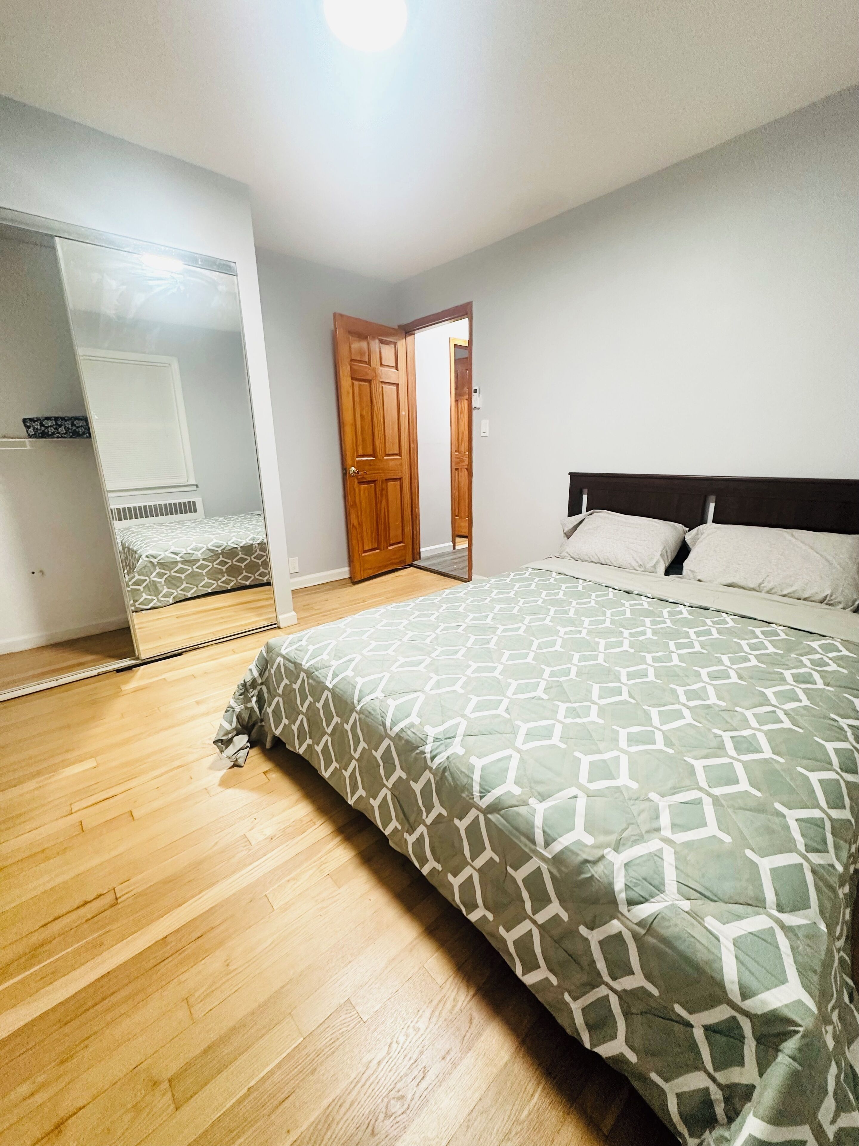 A bedroom with a wooden floor, a bed with a patterned bedspread, a mirror, and a door leading to another room.