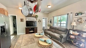 A spacious living room with a large window, a grey sofa, and a dining area with a table set for two.