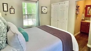 A bedroom with a bed, a window with curtains, a door, and a small bathroom visible through an open door.