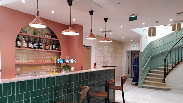 Eine Bar mit grüner Fliesenwand, einer Wein- und Spirituosenregal sowie Weidenbarhocker.