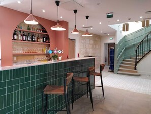 Zona de bar con un revoque verde de azulejos, una estantería para vinos y licores, y taburetes de bar de mimbre.