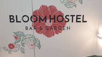A staircase with a wooden handrail and black balusters, leading to a room with a green wainscoting and a wall decorated with floral patterns and the text "BLOOMHOSTEL BAR & GARDEN".