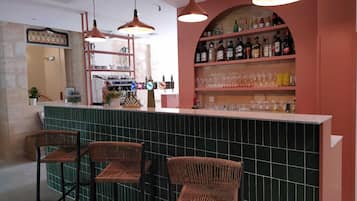 A bar area with a counter, three wicker chairs, a coffee machine, and shelves with bottles and glasses.