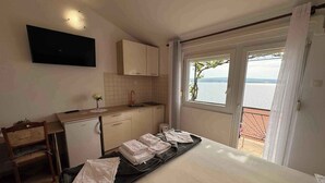 A hotel room with a bed, a small kitchenette, a TV mounted on the wall, and a balcony with a view of the sea.