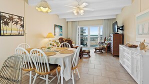A dining area with a table set for four, surrounded by white chairs, a ceiling fan, and a beach-themed painting on the wall.