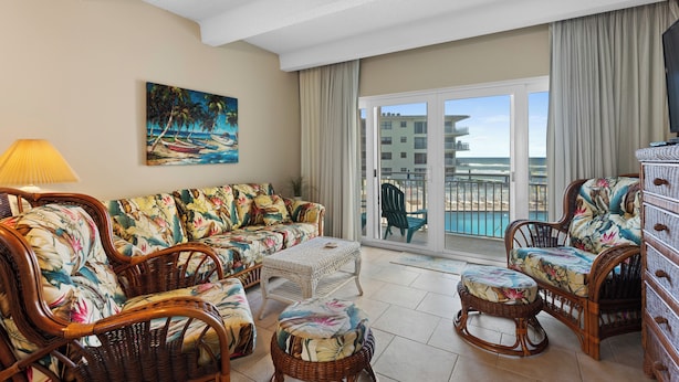 A living room with wicker furniture, a painting of a beach scene, and a view of a pool and buildings through a glass door.