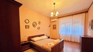 A bedroom with a wooden bed, a nightstand, a bedside table, a chandelier, a window with curtains, and a wall with framed pictures. - Holiday apartment first row to the sea with balcony (Crikvenica)