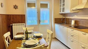 A dining area with a wooden table set for two, a window with curtains, a radiator, and a kitchen with white cabinets and a sink. - Holiday apartment first row to the sea with balcony (Crikvenica)
