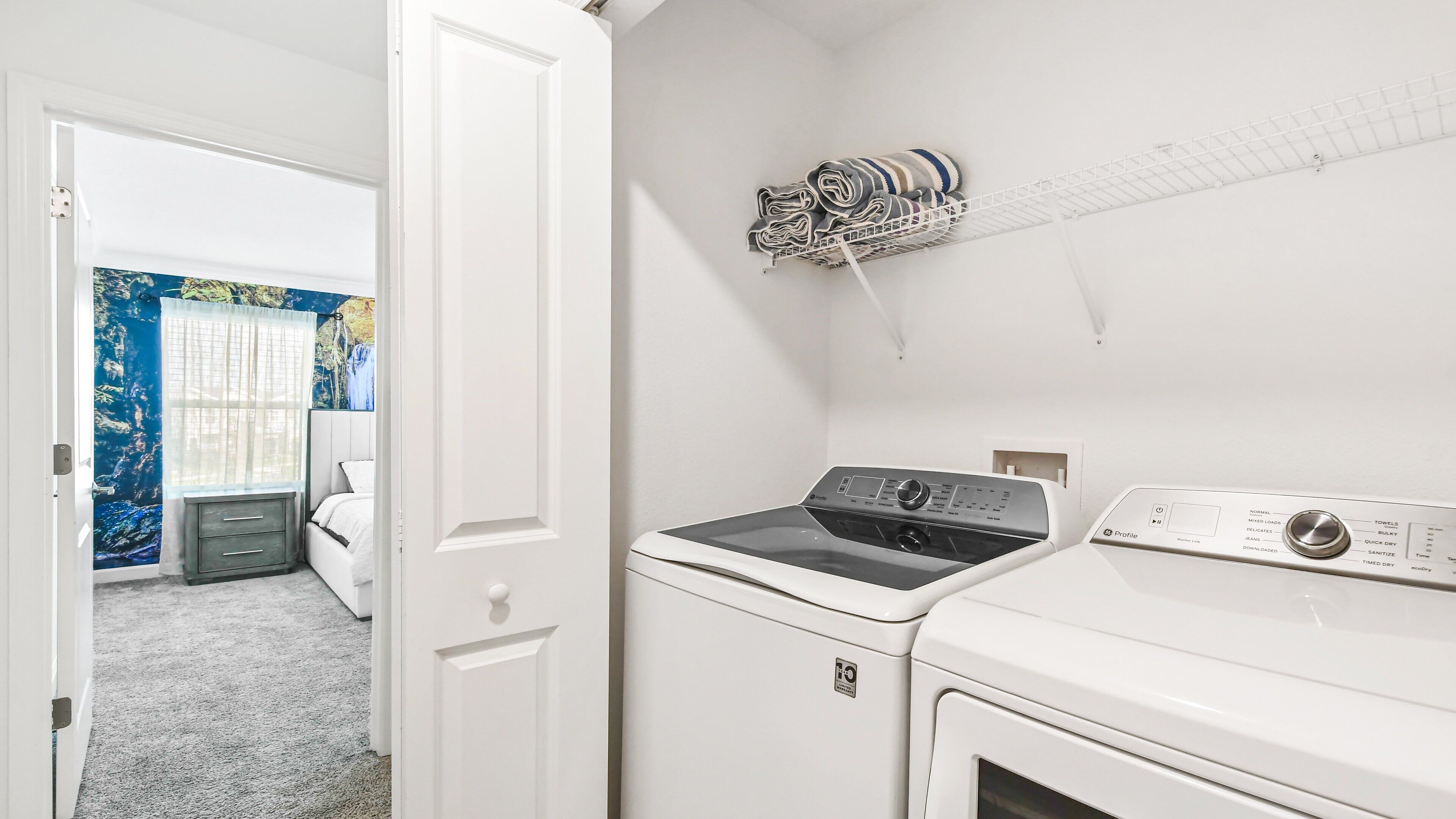 Un cuarto de lavado con lavadora y secadora de carga frontal, un tendedero con toallas dobladas, y una puerta que da a un dormitorio con cama y pared azul con una escena de naturaleza.