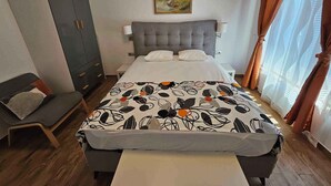 Une chambre d’hôtel avec un lit, une tête de lit grise, une table de chevet, une lampe, une armoire, une chaise et un tableau au mur.
