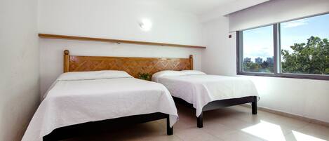A room with two beds, a wooden headboard, a large window, and a view of buildings and trees.