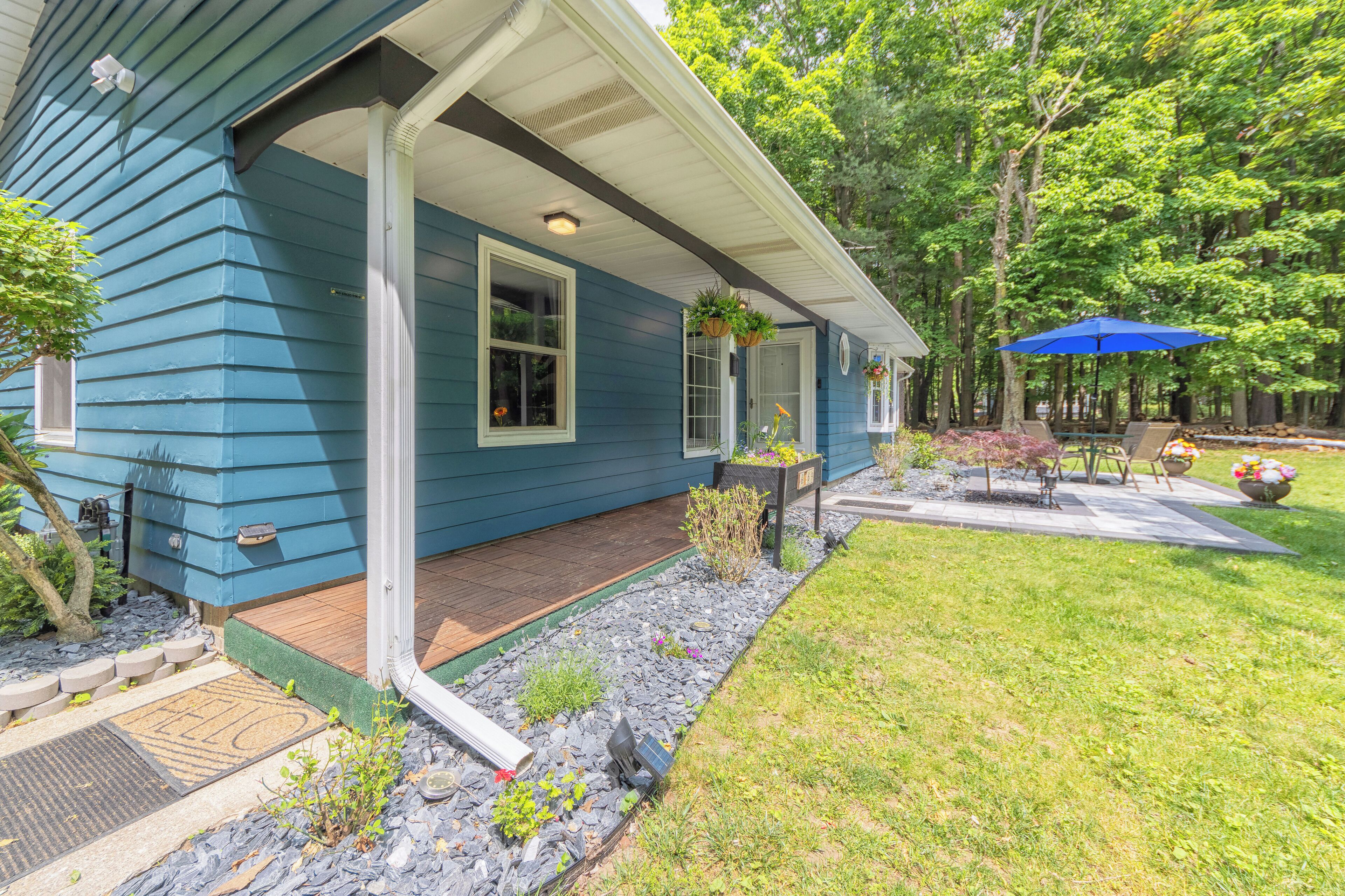 A blue house with a covered porch, a welcome mat, and a garden with a patio table and umbrella.