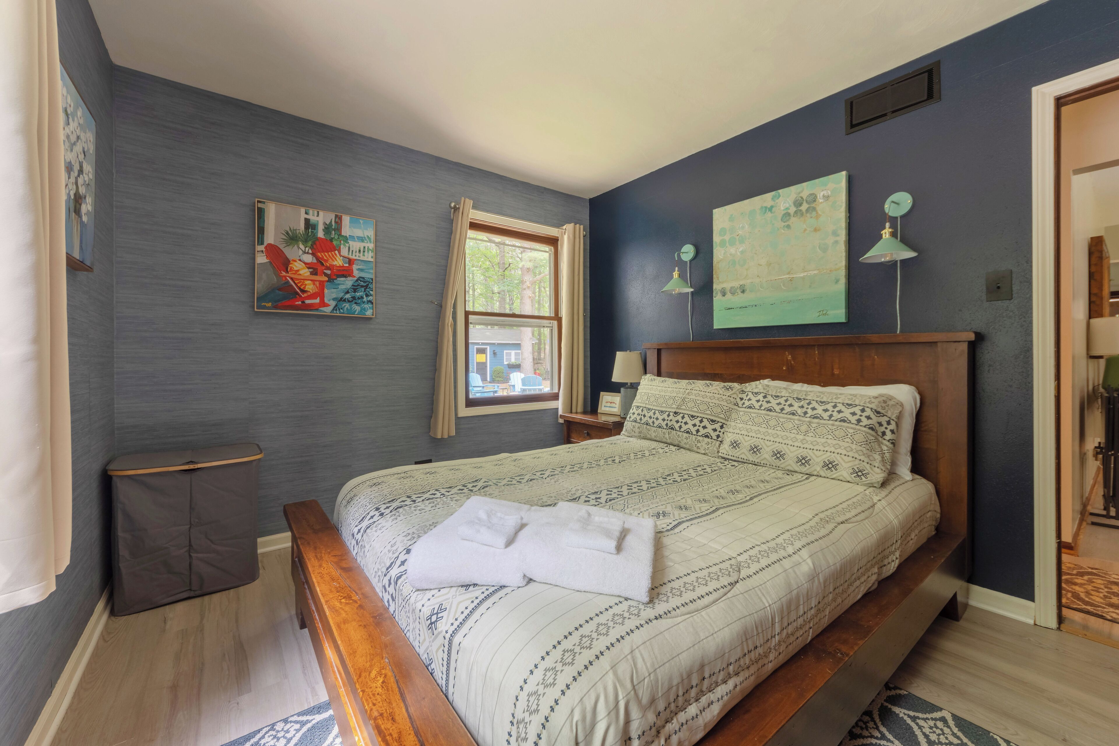 Una cama bien hecha con una colcha y almohadas estampadas, una mesita de noche con una lámpara, una ventana con cortinas y obras de arte en las paredes.