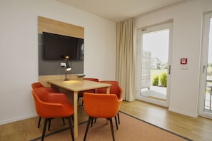 A modern room with a wooden table and orange chairs, a flat-screen TV mounted on the wall, and a sliding glass door leading to an outdoor area.