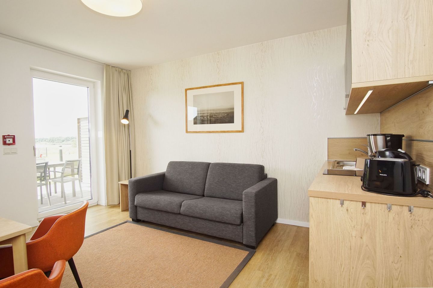Ein modernes Hotelzimmer mit grauem Sofa, einer kleinen Küchenzeile inklusive Kaffeemaschine und einem großen Fenster mit Blick ins Freie.