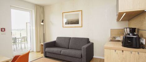 Ein modernes Hotelzimmer mit grauem Sofa, einer kleinen Küchenzeile inklusive Kaffeemaschine und einem großen Fenster mit Blick ins Freie.