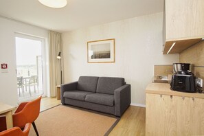 A modern hotel room with a grey sofa, a small kitchen area with a coffee maker, and a large window offering a view of the outdoors.