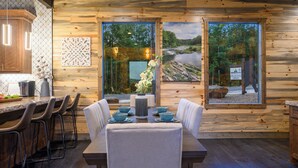 Ein Holzinterieur mit Essbereich, inklusive Tisch für vier Personen, Barhocker und einem großen Fenster mit Blick auf einen Fluss und Wald.