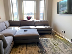 A living room with a sectional sofa, a flat-screen TV mounted on the wall, and a large window with a view of the outdoors.