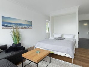 Ein Schlafzimmer mit einem Bett, einem Holztisch, einer schwarzen Couch, einem Blumentopf und einem Gemälde mit einer Strandansicht an der Wand.